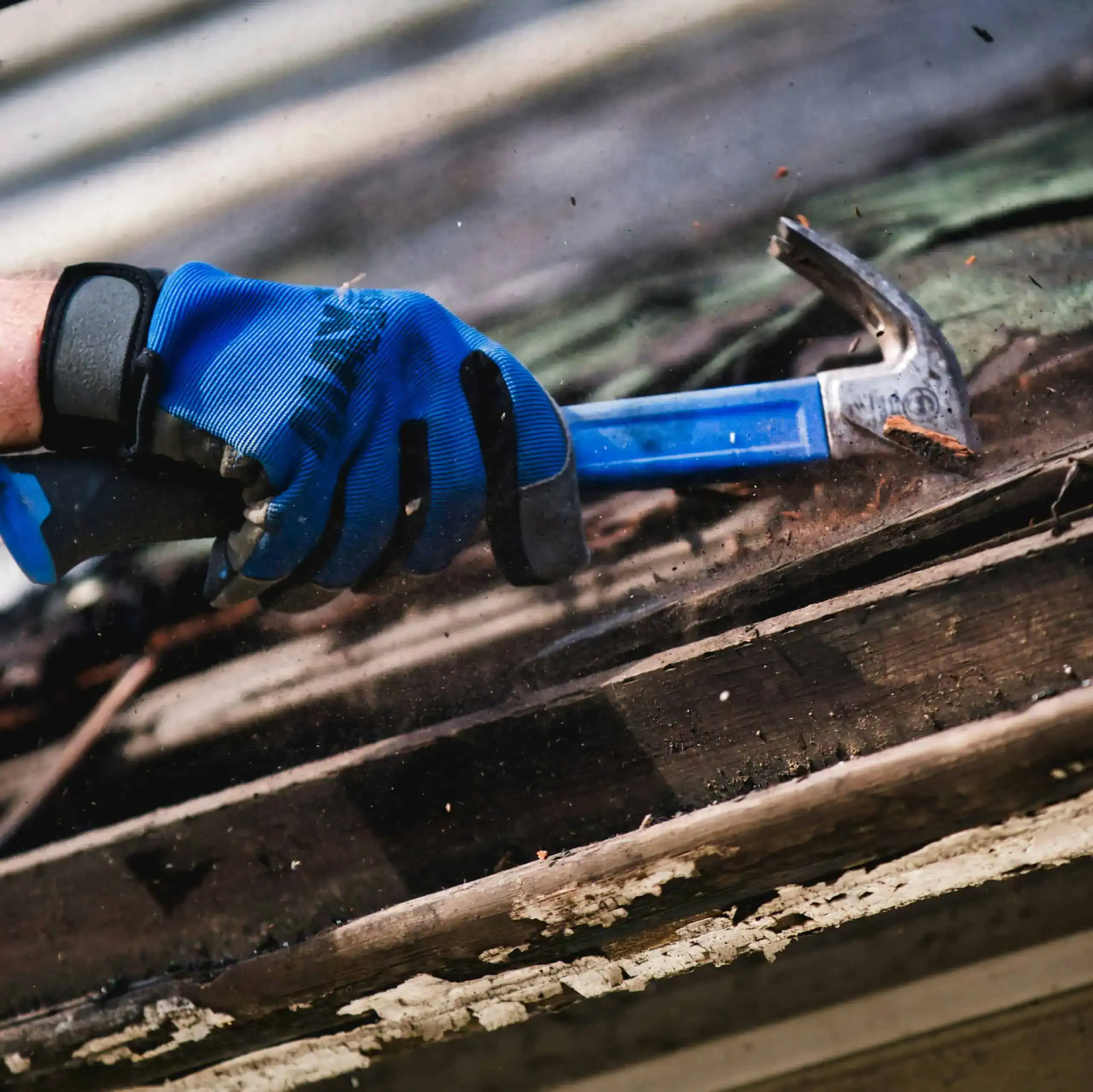 Close-up of a construction worker’s gloved hand using a hammer to remove old wooden roofing boards.