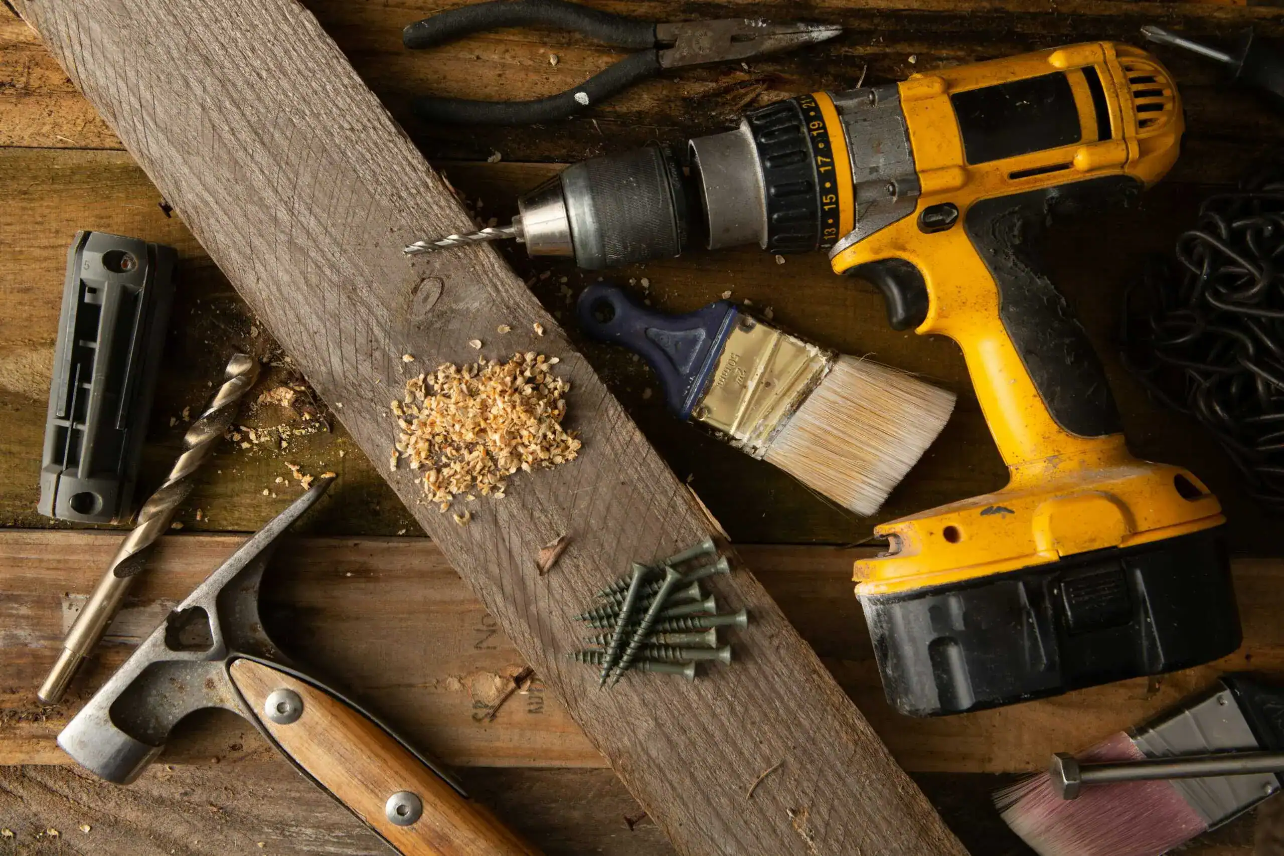 Assortment of woodworking tools including a cordless drill, hammer, screws, paintbrushes, and pliers on a wooden surface.